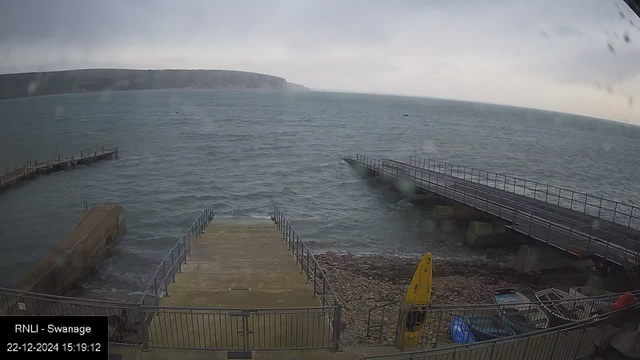 A webcam view of a cloudy seascape showing a pier extending into the water. In the foreground, there is a boat ramp leading into the sea, bordered by railings. Several small boats and a yellow kayak are visible on the rocky shore next to the ramp. The sea appears calm, and the horizon is a line of distant cliffs under a grey sky.