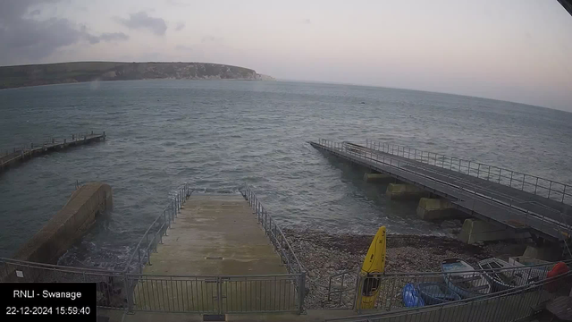 Webcam view of the sea at Swanage, showing choppy waves and a cloudy sky. In the foreground, there is a concrete slipway leading to the water, flanked by metal railings. To the right, a pier extends over the water. On the pebbly shore, several small boats and a yellow kayak are visible. The timestamp on the image reads "22-12-2024 15:59:40."