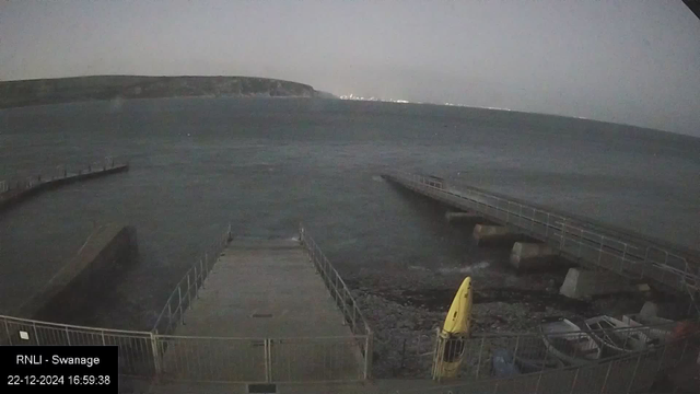 The image shows a view from a seaside webcam at dusk. Two piers extend into a vast body of water, creating an open corridor. In the foreground, there's a small landing area with safety railings and a yellow kayak placed upright. The horizon shows a distant landmass, and faint city lights are visible in the distance across the water. The bottom left corner has text that reads "RNLI - Swanage, 22-12-2024 16:59:38."