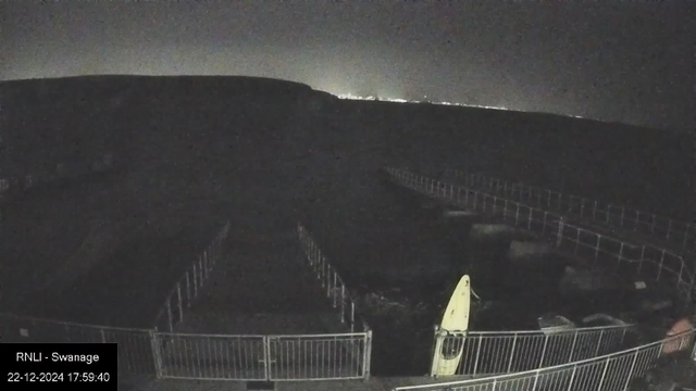 Nighttime webcam view of a fenced area leading towards dark hills or a cliff. The sky is dark with a faint glow on the horizon, possibly from distant lights. In the foreground, there is a yellow kayak or boat leaning against the fence. A timestamp in the bottom left corner reads "RNLI - Swanage 22-12-2024 17:59:40."