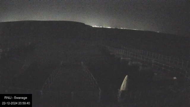 A dark, nighttime view from a webcam. The image shows a silhouette of a hill against a night sky with scattered lights in the distance. In the foreground, there are faint outlines of structures or barriers, and a vertical white shape is barely visible. The bottom left displays text: "RNLI - Swanage" and the timestamp "22-12-2024 20:59:40."