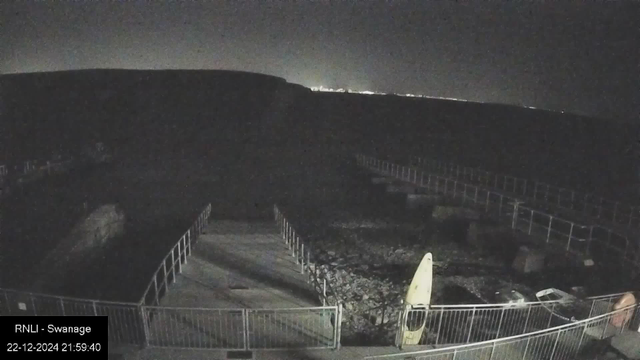 Nighttime view from a webcam showing a coastal scene. The foreground features a fenced pathway with a sign and a yellow surfboard resting against a railing on the right. The background is mostly dark, with faint silhouettes of cliffs and the distant glow of city lights just visible on the horizon. The image includes a timestamp at the bottom left corner that reads "22-12-2024 21:59:40" and "RNLI - Swanage."