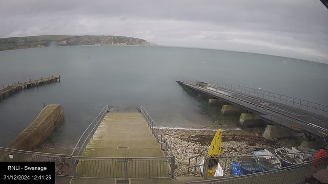 A cloudy sky looms over a calm sea, with a distant view of a hilly coastline. In the foreground, a concrete ramp leads down to the water between two metal fences. To the right, a pier extends over the water with railings along its edge. On the left side, there's a narrow dock. At the bottom right, several small boats and kayaks are stored on a rocky shoreline. The image has a timestamp from December 31, 2024, and is labeled "RNLI - Swanage".