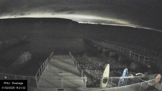 A nighttime view from a webcam showing a dimly lit pier or walkway with railings on both sides leading towards a body of water. The sky is dark with a faint glow on the horizon, possibly from city lights. In the foreground, there are a couple of kayaks or small boats beside the walkway. The image is labeled with "RNLI - Swanage" and a timestamp of February 1, 2025, at 18:21:02.