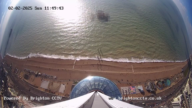 **Alternative Text:** A high-angle view of a sandy beach along the coastline, with gentle waves lapping at the shore. The image captures the reflection of sunlight glistening on the water's surface. In the background, the remains of a pier can be seen extending into the sea. To the right, there are various amusement attractions and structures along the boardwalk. The date and time display in the top left corner shows "02-02-2025 Sun 11:49:43." The bottom of the image includes the logo "Powered By Brighton CCTV" and the website URL.