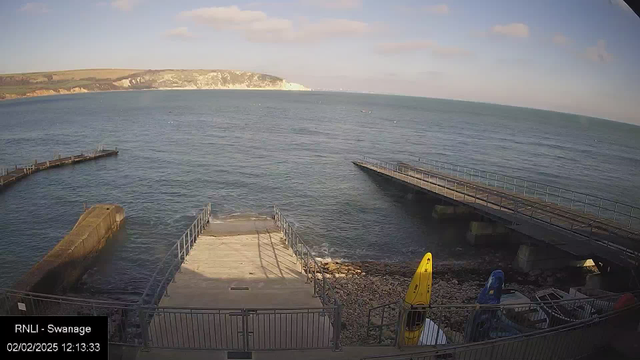 **Alternative Text:** A coastal scene viewed from a webcam. The image captures the sea with gentle waves under a clear blue sky. To the left, there is a concrete structure extending into the water, and a wooden pier is visible to the right. There are two seated kayaks in yellow and blue resting near the shore. The sandy beach area is minimal, bordered by rocks. In the distance, white cliffs rise along the shoreline. The timestamp indicates it is noon on February 2, 2025, from a site in Swanage.