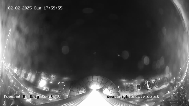 A black and white image from a webcam showing a view of a busy street at dusk. The center features a bright light source, possibly a building or structure illuminated from above. To the left, faint outlines of vehicles and lines of light suggest traffic. The lower part of the image displays indistinct shapes, likely buildings or other urban elements. The top of the image has a timestamp indicating the date and time.