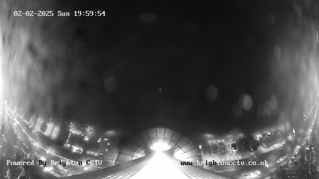 A black and white image captured by a webcam showing a night scene with a prominent bright light at the center, likely from a street or building. The lower part of the image features indistinct outlines of structures, possibly buildings or vehicles, with faint light reflections scattered throughout the scene. There are text indicators at the bottom left, stating "Powered By Brighton CCTV," and a website URL at the bottom right. The timestamp in the top left corner shows "02-02-2025 Sun 19:59:54."