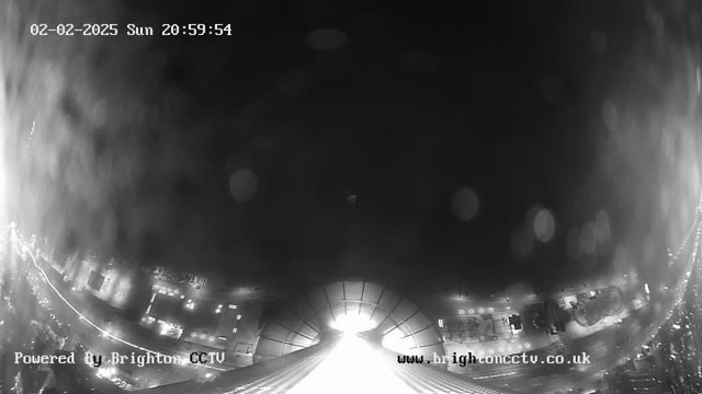 A black-and-white image captured by a webcam, showing a view of a cityscape at night. The scene includes a bright light source in the center, possibly a street or building illumination, while the edges of the image display faint outlines of buildings and streets illuminated by lights. There is a timestamp indicating the date and time at the top, and the bottom includes a watermark for the CCTV service provider. The image appears slightly blurred, likely due to moisture on the lens.