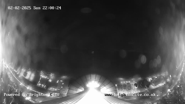 A black and white webcam image depicting a cityscape at night. The bottom of the image shows a well-lit area with outlines of buildings and street lights. The upper portion is darker with a few blurred spots, possibly caused by raindrops on the camera lens. The date and time are displayed at the top, and the image includes a watermark indicating it is powered by Brighton CCTV.