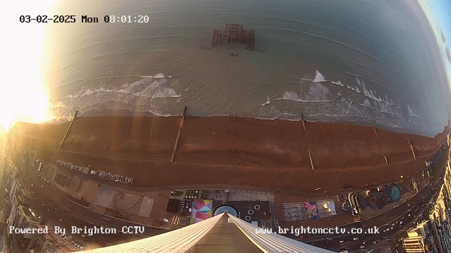 A panoramic view of a beach with waves rolling in, taken from an elevated position. The sun is rising, casting a bright light over the scene, creating a glare on the surface of the water. In the foreground, a wide sandy beach is visible, with several piers extending into the sea. People can be seen walking on the beach, and further back, there are colorful beachside attractions and structures, along with a road lined with buildings. The date and time display is shown in the top corner.