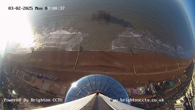 A view from above showing a sandy beach with gentle waves lapping at the shore. In the distance, a partially submerged pier structure is visible. Below, there are remnants of beach amenities and a promenade with several buildings. The image has a bright sun glare reflecting off the water, and the sky transitions from bright near the sun to a clearer blue further away. A central circular structure is prominent in the foreground. The time and date are displayed at the top of the image.