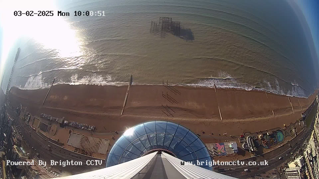 Bird's-eye view of a beach with a sandy shoreline stretching across the bottom. The sea is visible in shades of blue, with gentle waves lapping at the shore. A pier is seen extending out into the water, and a partially submerged structure is located further out. To the right, there are beach facilities and amusement park rides. The sky is bright with sunlight reflecting on the water, indicating a clear day. The image timestamp shows it is Monday, February 3, 2025, at 10:00:51.