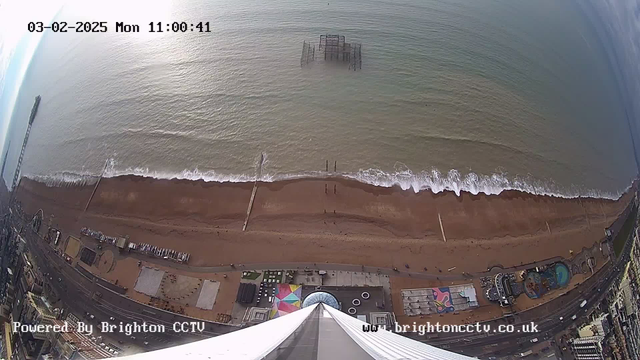 Aerial view of a beach and ocean on a cloudy day. The beachfront is sandy with some waves lapping at the shore. There is a wooden pier visible extending into the water, and part of a derelict structure partially submerged offshore. Below the beach, amusement rides and a colorful area can be seen, with some people walking in the sand. The image includes timestamps and a logo in the bottom corners.