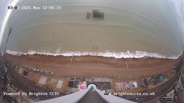 A panoramic view of a beach under a cloudy sky, taken from above. The image shows a sandy shoreline with gentle waves lapping at the edge. In the distance, a dilapidated pier extends into the water. There are various structures and attractions visible on the beach and along the promenade, including a circular pattern and some colorful designs. A few vehicles can be seen on the road parallel to the beach. The timestamp in the top left corner indicates the date and time of the image.