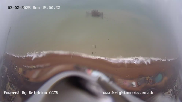 A view of a sandy beach with gentle waves lapping against the shore. In the distance, a pier is partially submerged in the water. The sky above is cloudy, and the time and date display shows "03-02-2025 Mon 15:00:22" at the top of the image. The bottom left corner contains the text "Powered By Brighton CCTV," while the website address is visible in the bottom right corner.