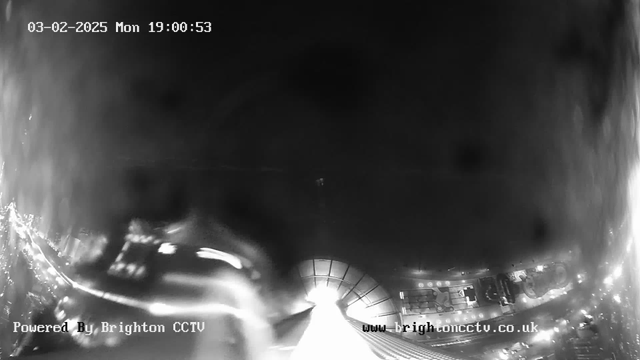 A black and white image from a webcam showing a nighttime cityscape. The upper half is dark with a few faint light sources. At the bottom, illuminated areas indicate city streets with some visible vehicles. The image is timestamped with the date and time displayed in the upper left corner, and branding for Brighton CCTV is present in the lower left and right corners. The overall visibility is low, giving a moody and atmospheric view of the scene.