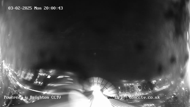 A blurry black and white image from a webcam, showing a partially obstructed view of a city scene at night. The foreground is distorted and unclear, while the background features light sources, suggesting buildings or streetlights. The date and time are displayed in the top left corner. The bottom part of the image includes text that indicates it is powered by Brighton CCTV.