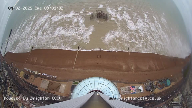 A wide-angle view of a beach and an ocean from a high vantage point. The sandy beach stretches along the bottom of the image, dotted with wooden groynes extending into the water. Waves crash gently against the shore. Further out in the water, remnants of a pier can be seen. The sky is cloudy, and there are patches of light on the water's surface. In the lower part of the image, various structures and a pool are visible along the promenade, with a circular building in the foreground. A timestamp indicates the image was taken on February 4, 2025, at 09:01.