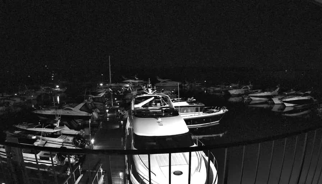 A nighttime view of a marina filled with boats. The scene is predominantly dark, with some boats illuminated by faint lights. A large white yacht is in the foreground, surrounded by smaller boats of various sizes and shapes, docked closely together. Reflections of the boats can be seen in the calm water. The surrounding area has limited visibility, with a few distant lights hinting at structures or dock areas.