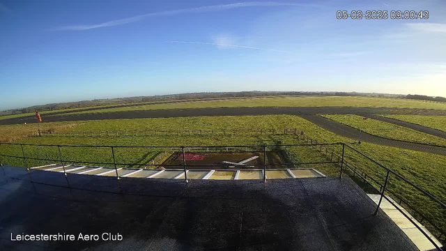 A clear blue sky with some wispy clouds is visible above a grassy airfield. In the foreground, a small airplane is parked on the tarmac, accompanied by a red and white windsock indicating the wind direction. The landscape includes a fenced grassy area and a winding taxiway leading off into the distance. The cam captures an expansive view of the airstrip and surrounding fields, typical of an airport setting.