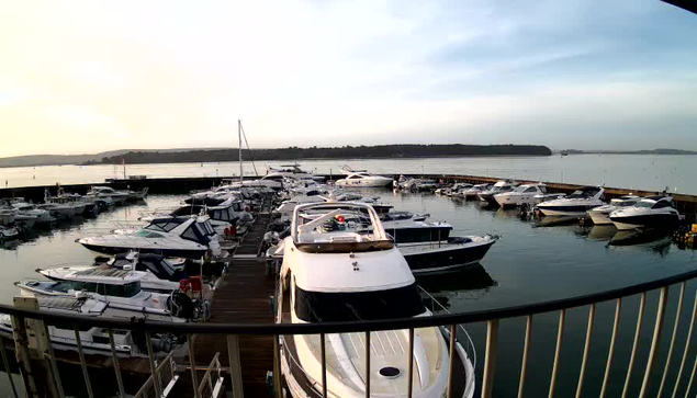 A view of a marina filled with various boats and yachts moored along a wooden dock. The scene features several white and blue vessels, with some cabins visible on the boats. In the background, a calm water surface reflects the sky, which transitions from a soft golden hue near the horizon to a pale blue. A distant landmass is visible across the water. The image is framed by a railing in the foreground.