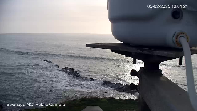 A view of the ocean captured from a webcam. In the foreground, there is a white container perched on a metal platform with a support structure. The surface of the water is calm with gentle waves, reflecting the light of the sun, which is low in the sky and creating a golden shimmer across the water. In the distance, a rocky shoreline can be seen extending into the water. The image is slightly hazy, suggesting a calm and serene atmosphere. The timestamp indicates the date and time of the capture.