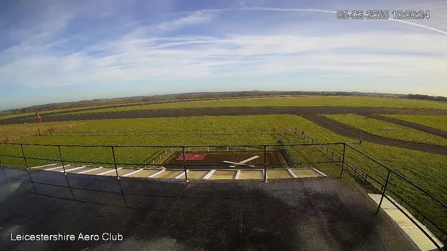 A wide view of an airport runway from an elevated position. The foreground features a railing, with the ground surface appearing dark and wet. Beyond the railing, a grassy area extends, bordered by a wooden fence. The runway is visible, marked with a large white "X." In the distance, open fields and a clear blue sky can be seen, with a few scattered clouds. The date and time are displayed in the upper right corner.