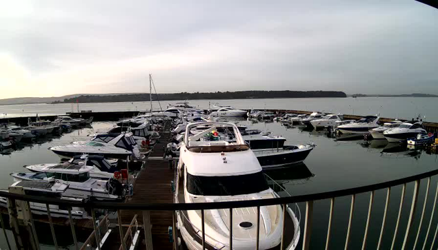 A marina filled with various boats docked along a wooden pier. Several motorboats and yachts are visible, many with different colors and designs. The water is calm and reflects the cloudy sky, with a distant shoreline and hills in the background. A railing in the foreground suggests a viewpoint from an elevated position.