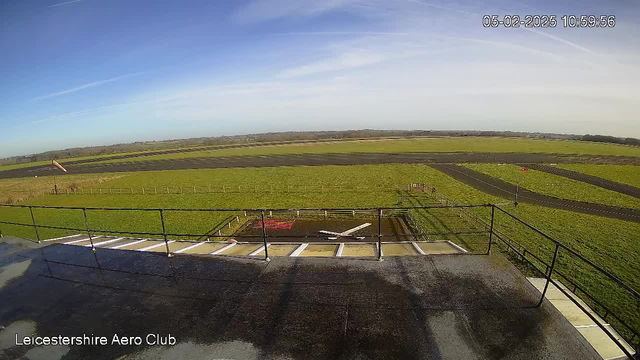 A clear sky is visible above a grassy field with a black runway. In the foreground, a view from a balcony shows a railing. Below, there is a large white X marking on the ground, suggesting a landing or takeoff area. To the left, a windsock is visible, indicating wind direction. The landscape extends into the distance with lines of trees and more fields. The date and time at the top indicate the image was taken on February 5th, 2025, at 10:59 AM.