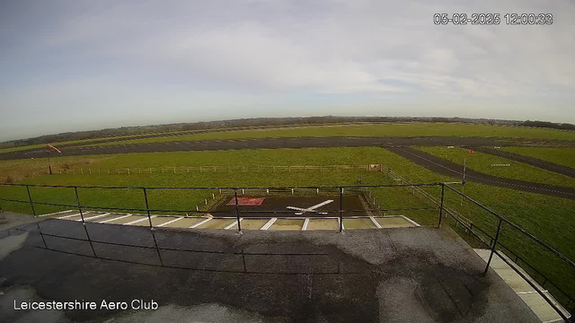 The image shows a landscape view from a webcam mounted on a building at the Leicestershire Aero Club. In the foreground, there is a flat rooftop with a safety railing. Below, a grassy area extends to a small runway that has a dark asphalt surface. There is a helipad with a white cross design, surrounded by a fence. In the distance, the horizon is visible with light clouds in a blue sky. A flag is seen at the edge of the runway, indicating wind direction.