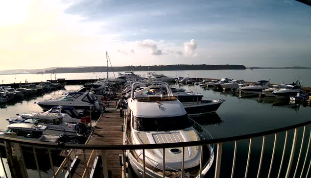 A busy marina scene showing several boats docked in a calm harbor. The image captures a wooden dock lined with various types of boats, including larger motorboats and smaller vessels. In the background, the water reflects a soft, cloudy sky with hints of sunlight. There are distant hills or land visible beyond the marina, and the scene has a peaceful, serene atmosphere.