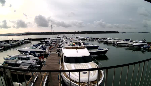 A panoramic view of a marina filled with various boats and yachts docked at a wooden pier. The water is calm, reflecting the cloudy sky above. In the background, there are trees lining the shore. The scene appears serene, with multiple boats of different sizes, some with colorful covers and flags. The atmosphere is overcast, suggesting a quiet, possibly breezy day by the water.