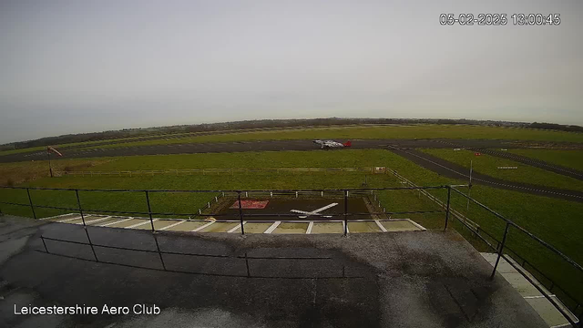 A view from a webcam at Leicestershire Aero Club, showing a grassy airfield under a cloudy sky. In the foreground, there is a railing, and a small section of the building roof is visible. On the runway, a small airplane is parked near a red area marked with a white X. Surrounding the runway are grassy fields and a fence, extending into the distance where additional runways and fields can be seen. The timestamp indicates the image was taken on February 5, 2025, at 1:00 PM.