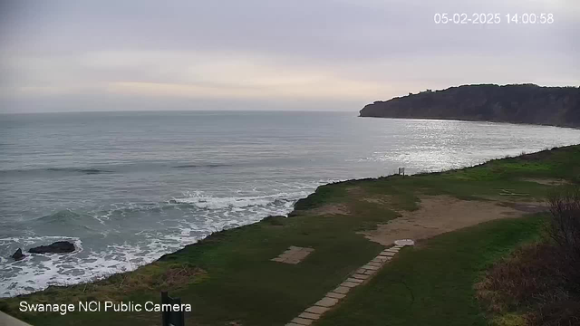 A coastal scene shows calm waters reflecting a pale sky, with waves gently lapping at the shoreline. The landscape features grassy areas and a sandy beach in the foreground, with a few rocks visible in the water. A distant cliff rises on the right side, under a cloudy sky. A timestamp at the top indicates the date and time of the image.