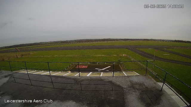 A view from a webcam at the Leicestershire Aero Club, showing a green grassy area adjacent to a runway. In the foreground, there is a white-painted area resembling a small airplane or helicopter landing spot. A fence surrounds the runway area, and a windsock is visible in the distance. The sky is overcast with gray clouds. The timestamp in the corner indicates the date and time as February 5, 2025, at 14:00:05.