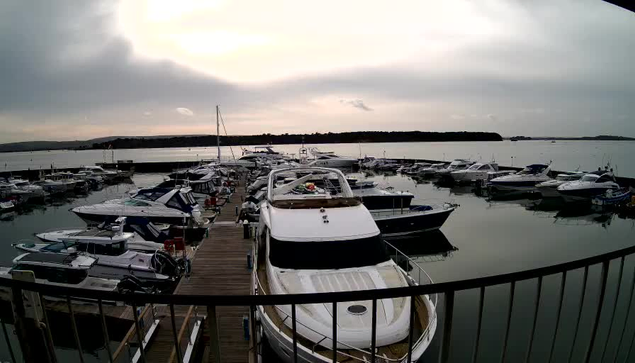 A view of a marina filled with numerous boats of various sizes docked along wooden piers. The scene is set under an overcast sky with soft gray clouds. In the foreground, a large white boat with a distinctive shape occupies a prominent position, alongside smaller boats with different colors and designs. The water is calm, reflecting the clouds overhead, while the distant shoreline is visible in the background.