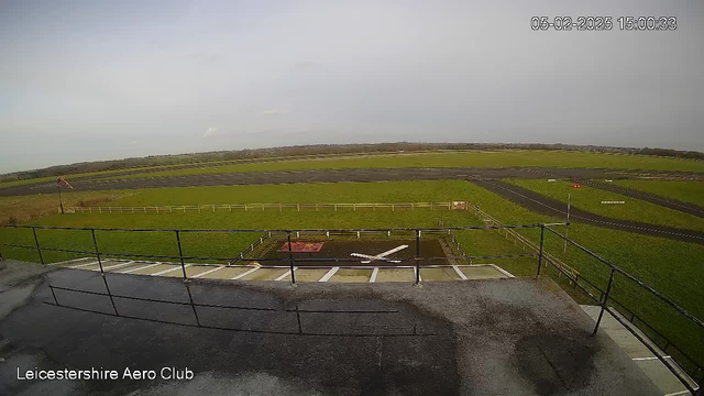 A view from a webcam positioned above a grassy airfield, with a light gray overcast sky. In the foreground, there is a railing and surface area with markings. The airstrip stretches out towards the horizon, flanked by patches of grass and dirt. A red windsock is visible to the left, indicating wind direction. In the background, a flat landscape stretches out, with trees lining the distance. The date and time are displayed in the top right corner.