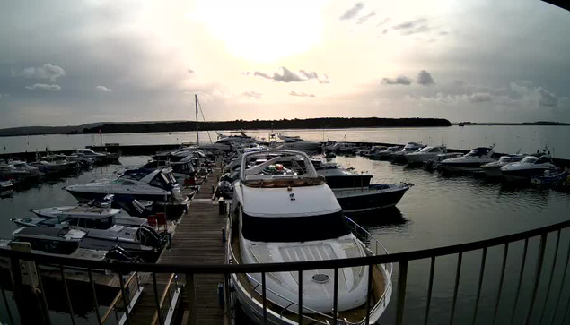 A view of a marina filled with various boats parked in the water. The foreground features a wooden dock leading out to the boats, with a large white yacht prominently visible. The scene is illuminated by soft, natural light with a cloudy sky overhead, reflecting off the calm water. In the background, a line of trees can be seen along the shoreline.