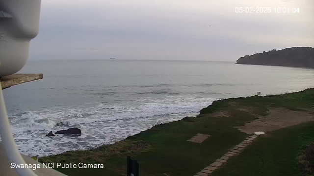 A coastal scene captured from a webcam. The foreground features grassy land with a narrow, paved pathway leading into the image. To the left, there are dark rocks partially submerged in the water. The ocean stretches across the middle of the frame, displaying gentle waves lapping against the shore. A ship can be seen in the distance, moving along a calm sea. The sky is overcast with soft, muted colors, indicating a cloudy day.