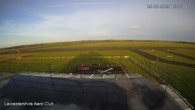 A panoramic view of an aerodrome on a clear day. In the foreground, there is a helipad marked with a red square and white cross, alongside a railing. The background features a vast expanse of green grass and a runway marked by lines, stretching toward the horizon. A windsock is visible on the left, indicating wind direction. The sky is mostly clear with a few clouds. The time and date are displayed in the top right corner.