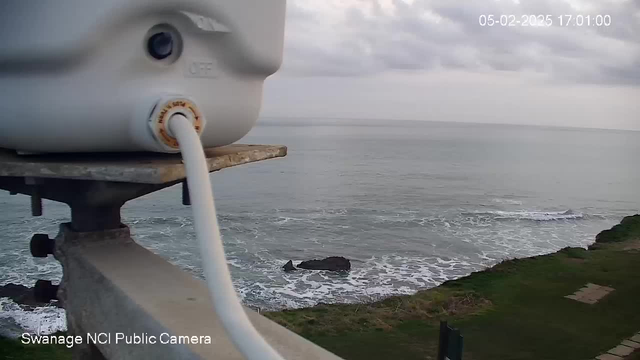 A gray webcam mounted on a railing overlooks the sea. In the foreground, a white cable extends from the camera. The background shows a calm ocean with gentle waves lapping against a rocky shoreline. The sky is mostly cloudy, and the coastline has patches of green grass. A timestamp in the top right corner indicates the date and time.