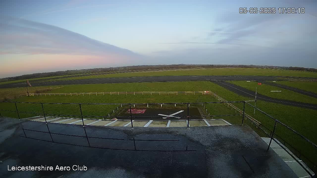 A wide view of an airstrip at the Leicestershire Aero Club. The foreground features a tarmac area with a painted white cross, surrounded by a grassy field and a wooden fence. In the background, the airstrip is visible, bordered by green fields and a clear sky transitioning from blue to soft pink hues at sunset.