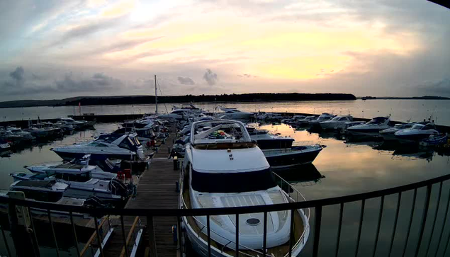 A serene marina scene at dusk features numerous boats docked in the water. In the foreground, a large white boat with a glossy finish is prominently positioned at the dock. Surrounding it are various smaller vessels of different shapes and colors, including blue and white boats. The water reflects the soft hues of the sunset, which includes shades of orange, pink, and purple, with a few clouds scattered across the sky. In the background, shoreline vegetation is visible against the horizon. The overall atmosphere is tranquil and picturesque.