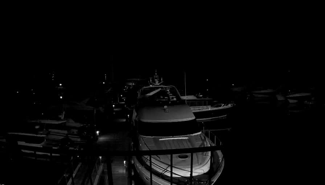 A dimly lit scene depicting several boats moored at a dock. The foreground features a white yacht partially illuminated, while multiple other boats are visible in the background, shrouded in darkness. Various small lights are scattered around the area, hinting at the presence of more vessels and the surrounding infrastructure. The overall atmosphere is quiet and calm, suggesting nighttime by the water.