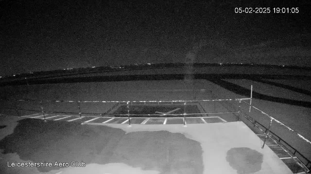 The image shows a dark scene from a webcam at the Leicestershire Aero Club. The foreground features a flat, empty surface with some markings and a railing along the edge. In the background, there is a vast area of dark land, likely an airstrip, stretching towards a dark sky. A few distant lights are faintly visible at the horizon. The overall atmosphere is quiet and still, indicating it is nighttime.