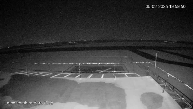 A dark, nighttime scene viewed from a webcam at Leicestershire Aero Club. The image shows a flat rooftop with a striped boundary near the lower edge. There are dark patches on the surface, possibly wet areas from rain. In the background, faint lights from a distant area are visible, indicating a horizon. The sky is black, and no aircraft or other structures are in sight. The time displayed is 19:59 on February 5, 2025.