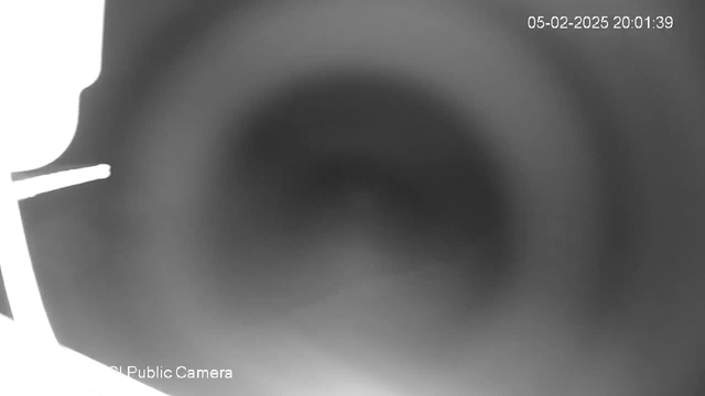 A blurred, grayscale image with a dark circular area in the center, suggesting a tunnel or a similar deep space. The left side of the image shows a vague outline of a figure, possibly a silhouette. The top right corner displays a timestamp indicating the date and time. At the bottom left, there is a label indicating this is from a public camera.