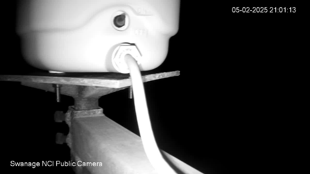 A close-up view of a white cylindrical container positioned on a dark surface. The container has a spout and an electrical cable connecting to it. The background is mostly black, indicating low light conditions, with some faint outlines of the surrounding area visible. The image is timestamped at 21:01:13 on February 5, 2025.