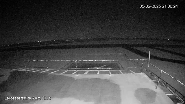 A dark nighttime scene captured from a webcam, showing an empty runway at Leicestershire Aero Club. The foreground features a flat surface with markings, and a railing along the edge. In the background, faint lights can be seen in the distance, likely from surrounding areas. The sky is mostly black, indicating it is night. The timestamp in the upper right corner reads February 5, 2025, at 21:00:24.